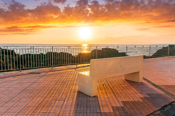 beautiful white stone bench on seafront embarkment during beautiful sunrise or sunset with sidewalk with pavement, sea surf and nice cloudy sunset sky