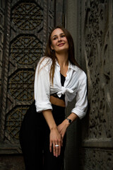 portrait of a beautiful brunette woman with long hair staying near wooden carved door in white shirt and black trousers 