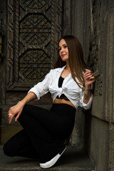portrait of a beautiful brunette woman with long hair sitting near wooden carved door in white shirt and black trousers 