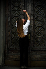 portrait of a beautiful brunette woman with long hair staying back near wooden carved door in white shirt and black trousers 