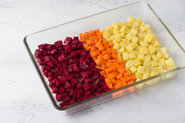 Raw diced vegetables: beets, carrots, potatoes seasoned with spices and vegetable oil in a glass baking dish on a light gray background