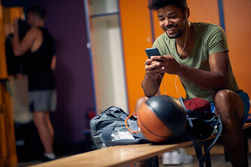 Joyful young man sitting in gym dressing room, looking at smartphone, preparing for workout.