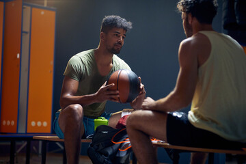 Two handsome young basketball players sitting in dressing room on bench across each other having a conversation, after workout.