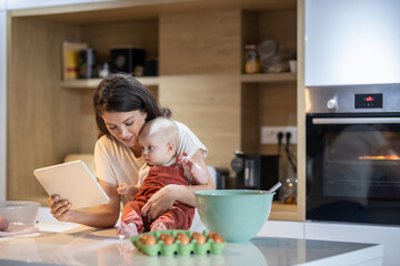 Young mom shows her baby something on her tablet as she holds her on the kitchen counter