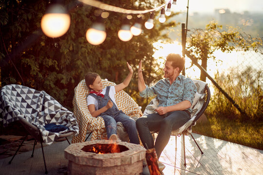Father and daughter high fiving, sitting outdoors in their yard by a fireplace enjoying time together, roasting smores during summertime.