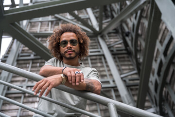 Portrait of a young man with afro hair wearing sunglasses on the stairs in the city, urban fashion...