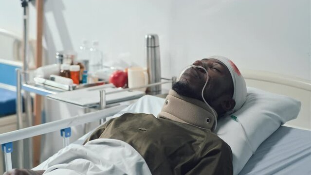 Medium close-up of African American wounded soldier lying alone on bed in hospital with closed eyes and bandage on head