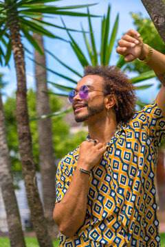 Portrait of afro haired man on summer vacation next to some palm trees by the beach smiling looking to the left leaning on a palm tree. Travel and tourism concept