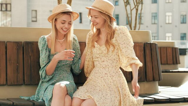 Two young beautiful smiling hipster female in trendy summer sundress and straw hats. Sexy carefree women in street. Positive models having fun, sitting at the bench and communicating, speaking 