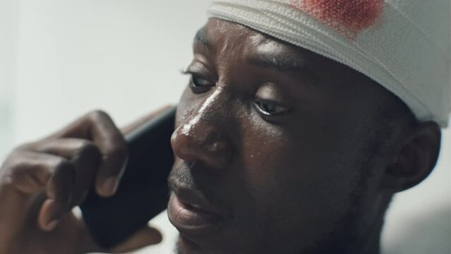 Close-up shot of African American soldier with bleeding head wound talking on phone with friend while lying in military hospital