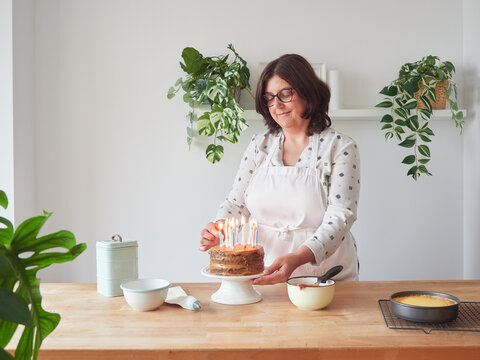 smiling woman placing lighted candles on chocolate cream birthday cake at home on wooden countertop with other decorating utensils
