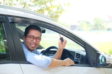 Adult Asian man smiling from inside his car while showing the car key - obrazy, fototapety, plakaty