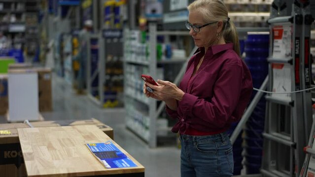 Mature woman uses phone to take photo of workbench furniture while shopping in large hardware store for home renovation project.