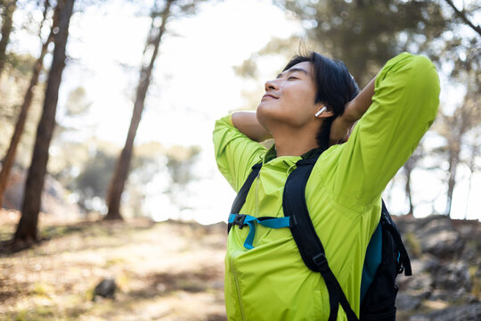 A Young Korean Man Is Posing With His Arms Behind His Head While His Eyes Are Closed. Concept Of Breathing Fresh Air In The Mountains, Disconnection From The City, Feeling Nature.