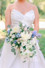 A beautiful bride in a white wedding dress holding a gorgeous bouquet of wildflowers in delicate shades of white, ivory, blue, and purple blooms and green leaves.