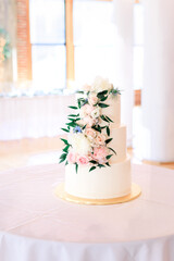 A white wedding cake is decorated with white and pink flowers and green leaves cascading down its three layers. 