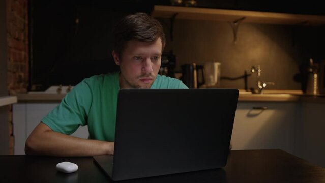 Home office concept. A man sits in the kitchen at night and works on a laptop