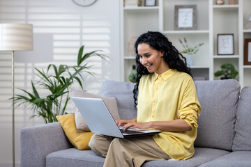 Young woman working with laptop sitting on sofa at home, successful Latin American woman in living room typing on computer keyboard working online remotely.