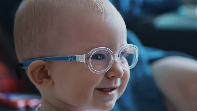 Happy smiling one year old baby with glasses is watching TV. White caucasian infant has poor eyesight or bad vision. Child kid has an eye disease, strabismus. Closeup, portrait