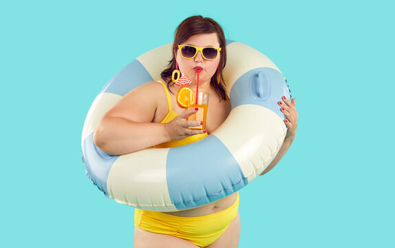 Portrait Of Funny Fat Plump Chubby Young Woman Wearing Yellow Holiday Swimsuit, Sunglasses And Earrings Standing On Blue Background, Holding Striped Beach Ring, And Drinking Fruit Cocktail From Glass