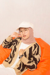 closeup of beautiful young woman in glasses and hat in white negligee and brown sweater sitting on orange bean bag with piercing eyes, on white studio background