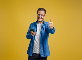 Portrait of smiling entrepreneur showing thumbs up and messaging on smartphone on yellow background