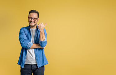 Fototapeta Portrait of smiling businessman pointing thumb at copy space for marketing over yellow background obraz