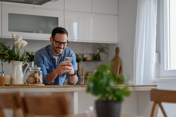Smiling young businessman sending messages over smart phone while standing at kitchen counter