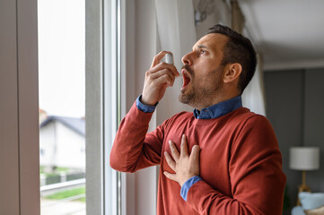 Asthmatic young man touching chest in pain and inhaling through asthma pump by window at home - obrazy, fototapety, plakaty