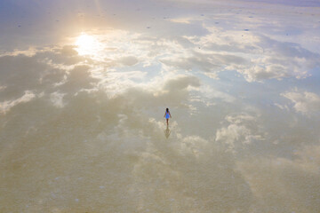 A woman in a blue dress walks along the shore of a salt lake with mirrored sky and clouds at sunset