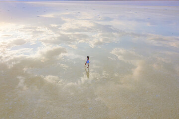 A girl in a blue dress walks along the shore of a salt lake with mirrored sky and clouds at sunset