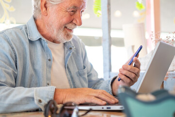 Smart work concept. Smiling senior man in coffee shop while working on laptop talking on phone....