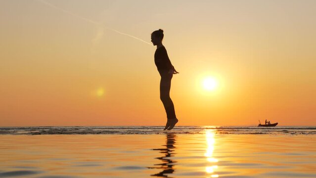 Young graceful gymnast girl make C Jump, vibrant sky and bright sun on background. Silhouetted shot of professional athlete at sea beach. Shallow water on foreground, girl stay at sand bank