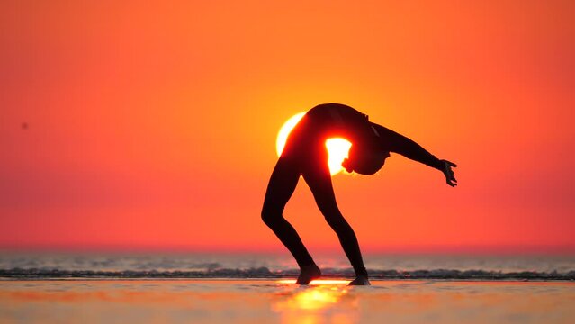 In breathtaking sunset setting, graceful gymnast girl bends backward, her slender silhouette standing out against vibrant sky and radiant sun. The telephoto perspective beautifully captures this stunt