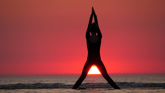 Slow exercise of young athlete, young girl slowly move hands up and spread out. She stay against sunset at sea shore, telephoto perspective, silhouetted shot