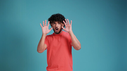 young frightened Hispanic or Arab man , raising hands with fear face in coral t-shirt on blue studio background. Human emotions, mood, psychological  concept