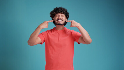 Happy confident swarthy man with toothy smile pointing with fingers on his mouth looking at camera isolated on blue studio background. Health care, dental treatment concept.