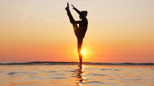 Witness determined spirit of young gymnast at sea beach as she attempts standing front split. Silhouetted against sunset, she gives her best, even though she experiences momentary loss of balance