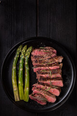 Pieces of Ribeye steak with asparagus on a black plate. View from above.