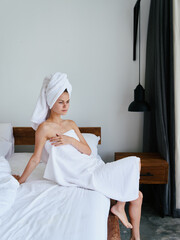 A woman sits on the bed after a shower with wet hair with a towel on her head in the bedroom and looks out the window, lifestyle