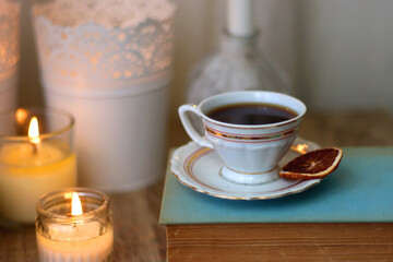 Cup of tea or coffee, plate of cookies, books, e-reader, pencil and lit candles on the table. Selective focus.