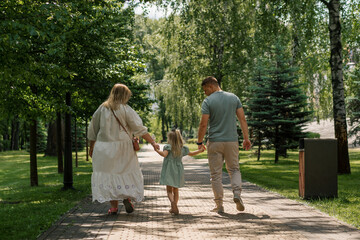 Fototapeta premium Little cute girl in green summer dress with parents in city park on a walk summer family lifestyle Childhood happy family rear view
