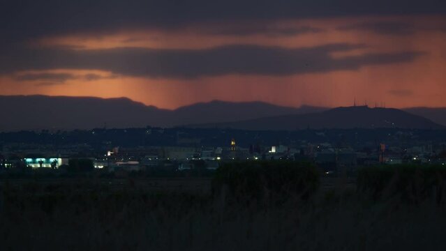 Against the backdrop of a red sunset sky and distant mountains, small houses of a suburban town and passing cars on the highway. Rain in the mountains
