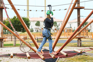 Teenage teen girl in climbing harness equipment, green sports safety helmet. Rope amusement park....