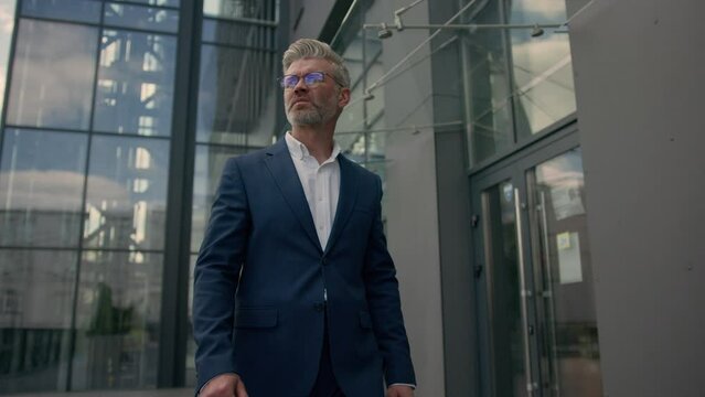 Relaxed middle-aged man senior mature business employer businessman walking in city outdoors near office building walk slowly check time looking at wrist watch shock lateness hurry go faster quickly
