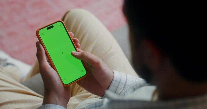 A man in casual clothes scroll the green screen of a mobile phone while sitting on the couch at home. Top view, no face, close-up of a man's hands