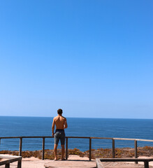 Young man traveler contemplating in a peaceful place in front of the ocean viewed from the back. Portugal Solo traveler.