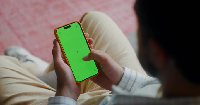 A man in casual clothes scroll the green screen of a mobile phone while sitting on the couch at home. Top view, no face, close-up of a man's hands