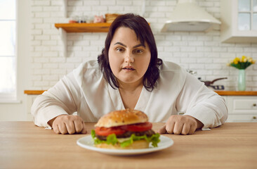 Young fat overweight excited woman sitting at the table in kitchen at home looking at the plate with big burger with a great temptation to eat it. Diet restrictions, losing weight concept.
