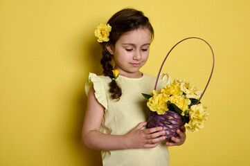 Obraz na płótnie Canvas Cute little kid girl with rose flowers in hairstyle, dressed in yellow summer dress, holding a purple wicker basket full of fresh yellow roses, isolated color studio background. Happy Children's Day